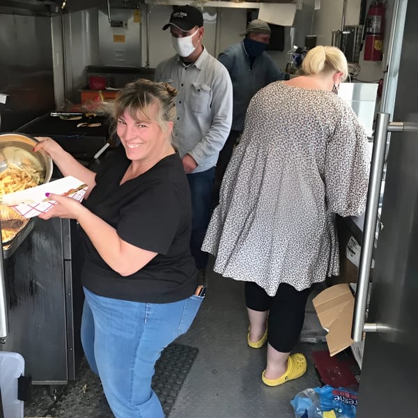 A smiling woman serves fries from a food truck kitchen, with other staff working behind her.