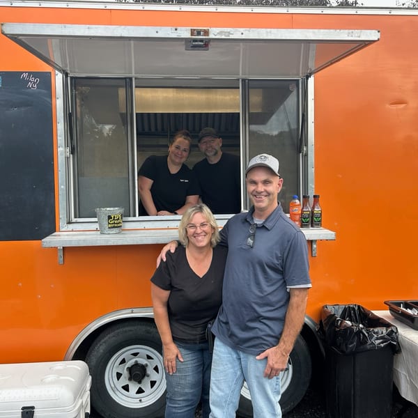 Four smiling Rustic Tables staff members, two inside and two outside, pose in front of an orange food truck.