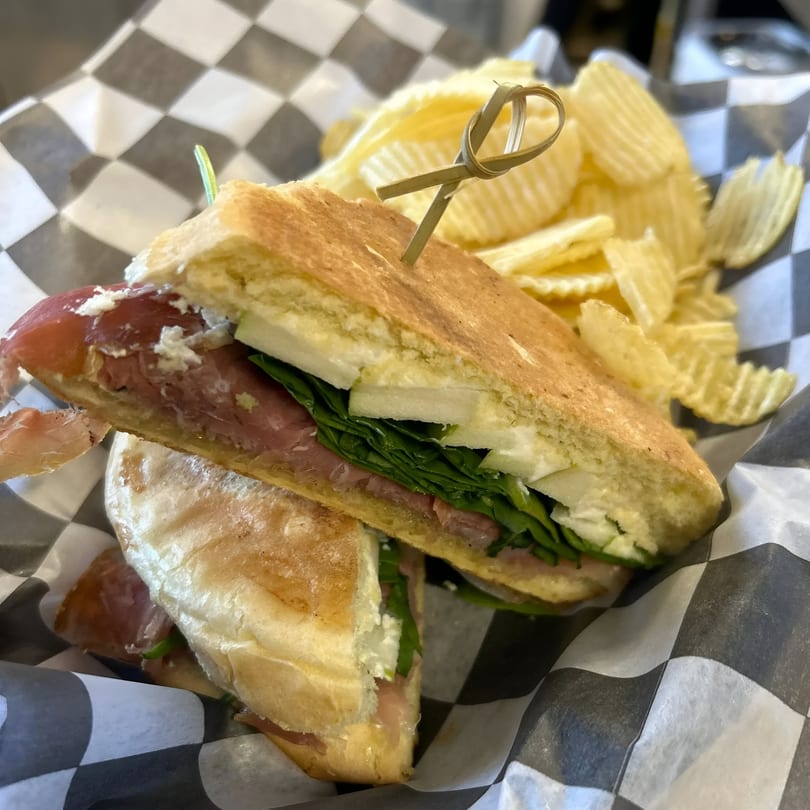 A Rustic Tables panini with meat, cheese, and greens, served with crinkle-cut chips.