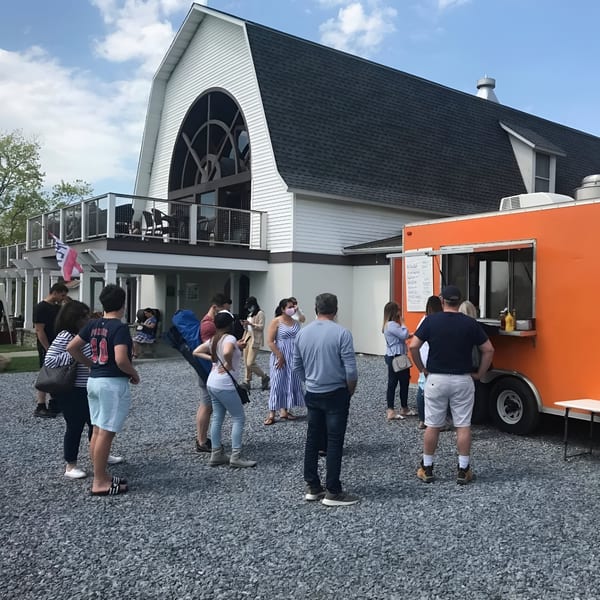 People line up at an orange food truck outside Rustic Tables Restaurant & Bar, a white barn-like building.