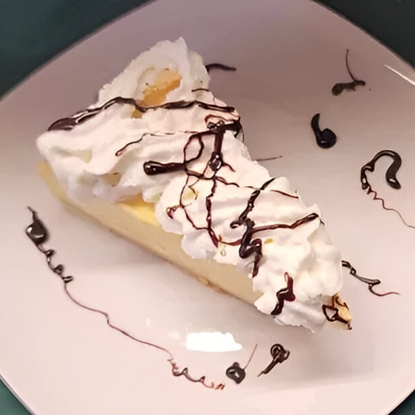Slice of cheesecake topped with whipped cream and chocolate drizzle on a white plate.