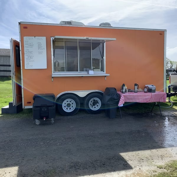 An orange food truck, "Cider Bros" menu visible, with a red and white checkered table outside.