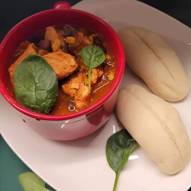 A red bowl of chili with chicken, beans, and spinach, served with two bread rolls.