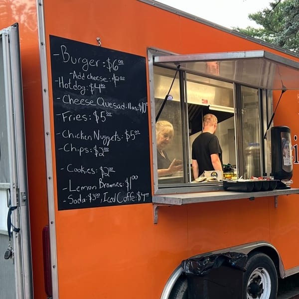 Orange food truck with a chalkboard menu and two people working inside the service window.