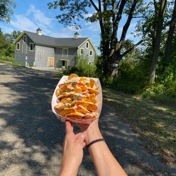Hands holding a tray of loaded potato wedges with a rustic barn in the background.