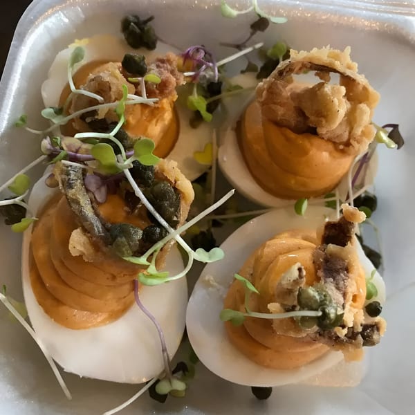 Four deviled eggs topped with fried onions, capers, and microgreens in a white container.