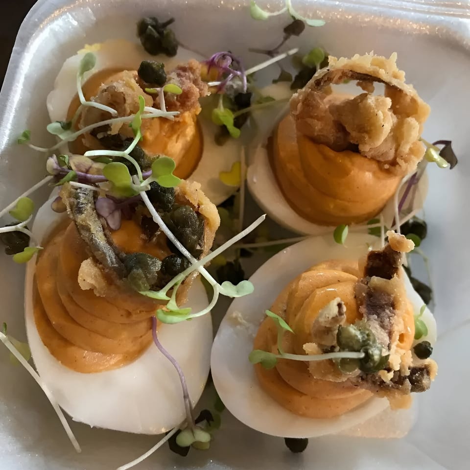 Four deviled eggs topped with fried onions, capers, and microgreens in a white container.