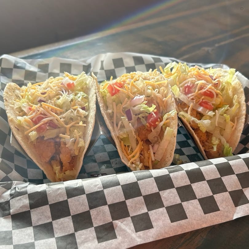 Three crispy tacos filled with chicken, lettuce, shredded cheese, and diced tomatoes in a checkered basket.
