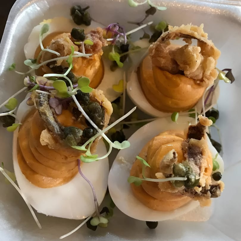 Four deviled eggs topped with fried anchovies, capers, and microgreens in a white dish.