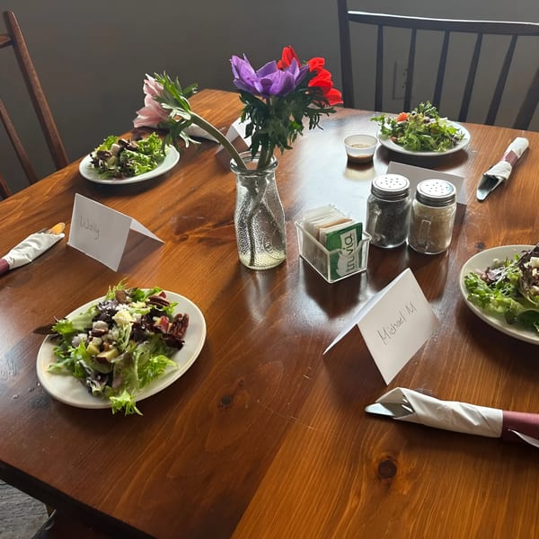 A rustic wooden table set with salads, place cards, silverware, and a vase of colorful flowers.