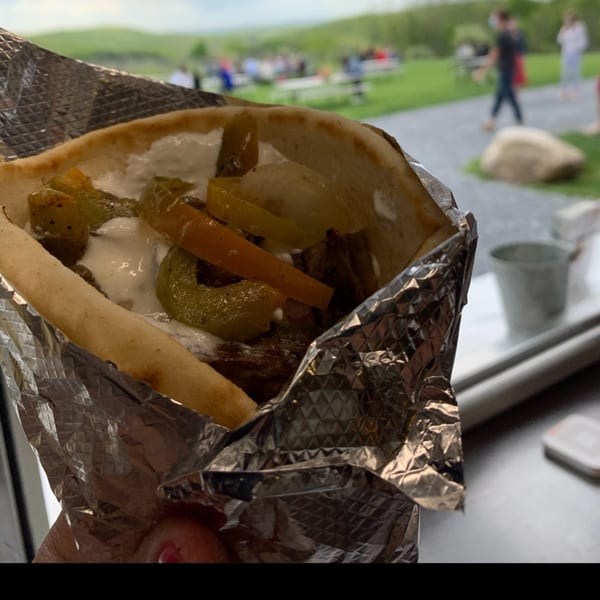 A foil-wrapped gyro with meat, white sauce, and peppers, with an outdoor picnic area in the background.