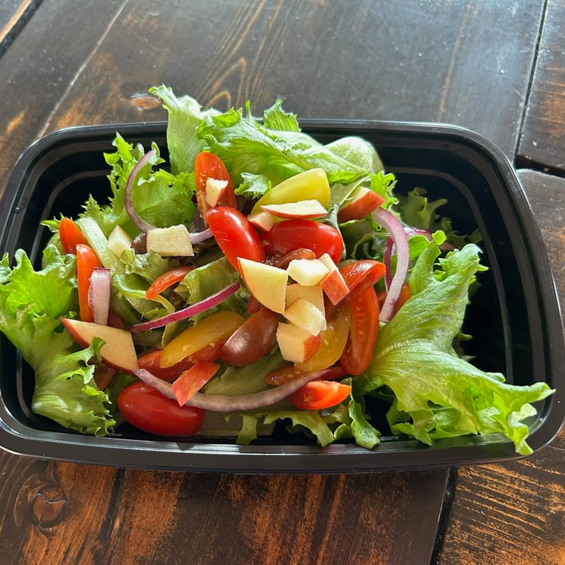 Fresh green salad with cherry tomatoes, red onion, and apple slices in a black to-go container.
