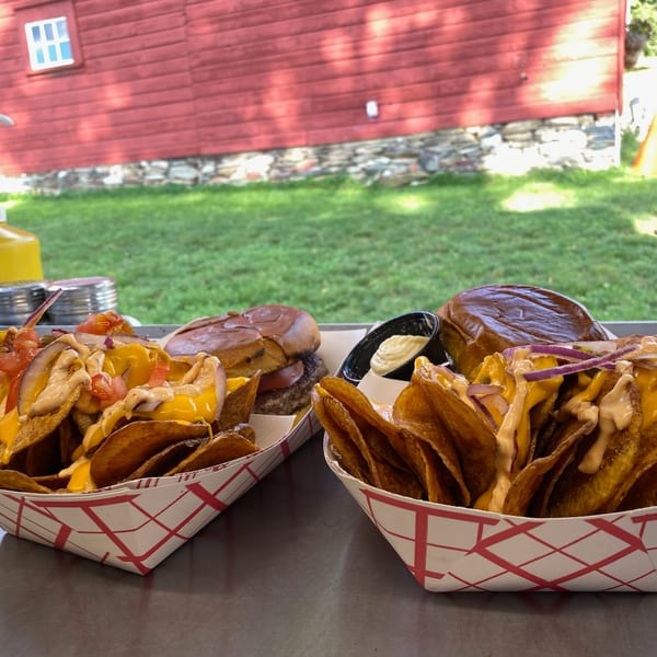 Two baskets of Rustic Tables' loaded potato chips and burgers on a metal table outdoors.